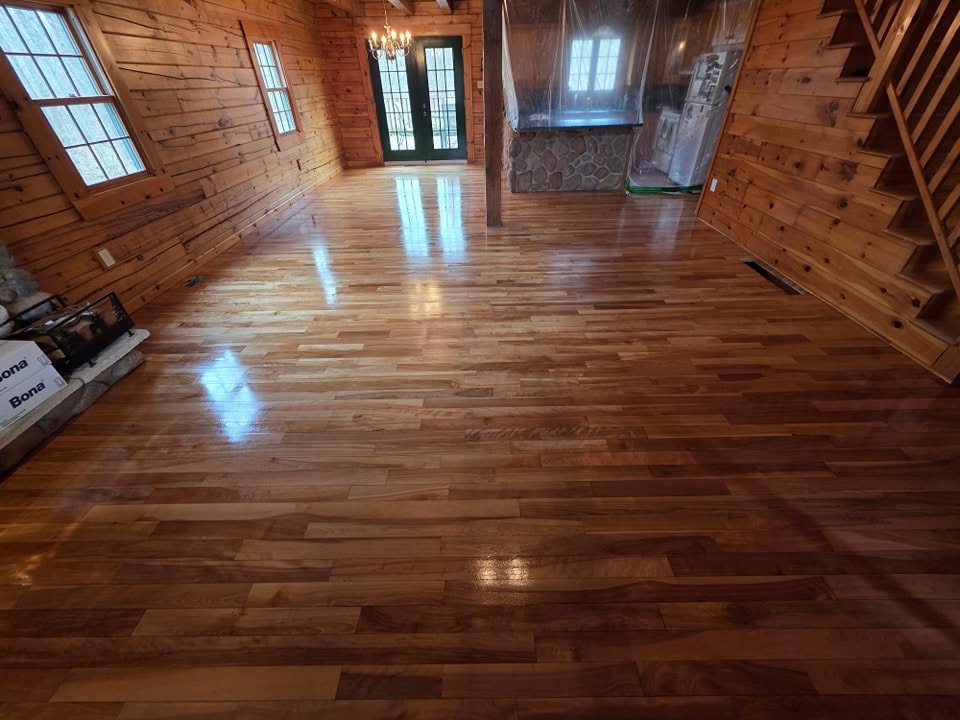 A wide hickory hardwood floor in a log home, freshly refinished to a deep gloss, with Bona finish boxes stacked at the edge of the room.