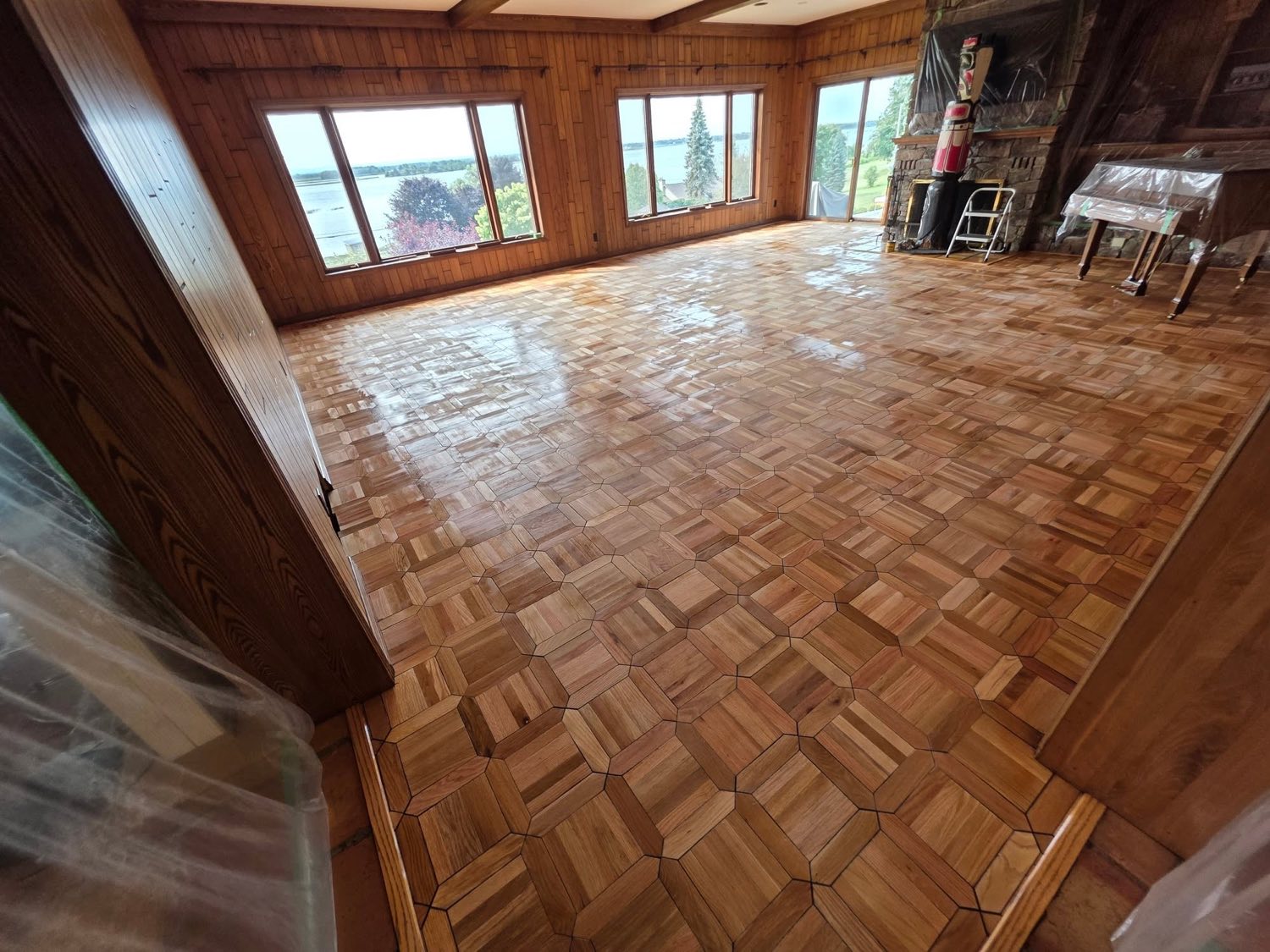 A large lakeside room with cedar walls and a stone fireplace, its intricate hexagonal parquet floor freshly refinished and reflecting light from a wall of windows.